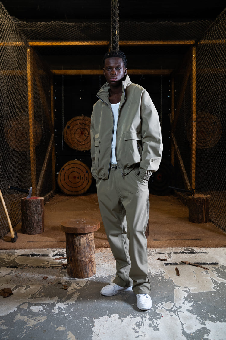 Frontansicht eines männlichen Models in einem lässigen, urbanen Outfit bestehend aus einer sandfarbenen "City Hunter" Jacke und Trackpants. Der relaxte Look wird durch weiße Sneakers abgerundet.