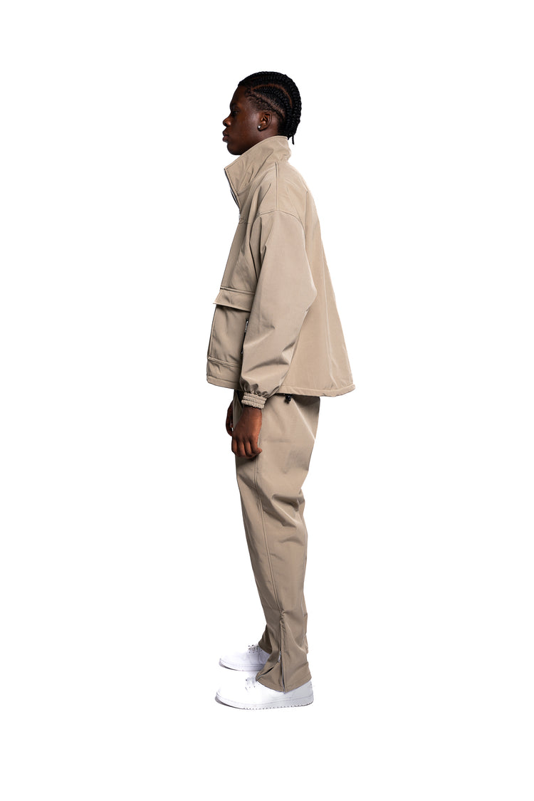 Seitliche Ansicht eines männlichen Models in einer vollständigen Streetwear-Kombination, bestehend aus einer sandfarbenen Jacke und passenden Hosen. Die minimalistische, einfarbige Aufmachung betont einen sauberen und modernen Look.