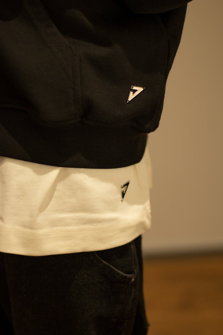 Detailansicht des Logo Metall Logo Emblems über dem Bund des schwarzen Pullovers sowie des weißen T-Shirts darunter.