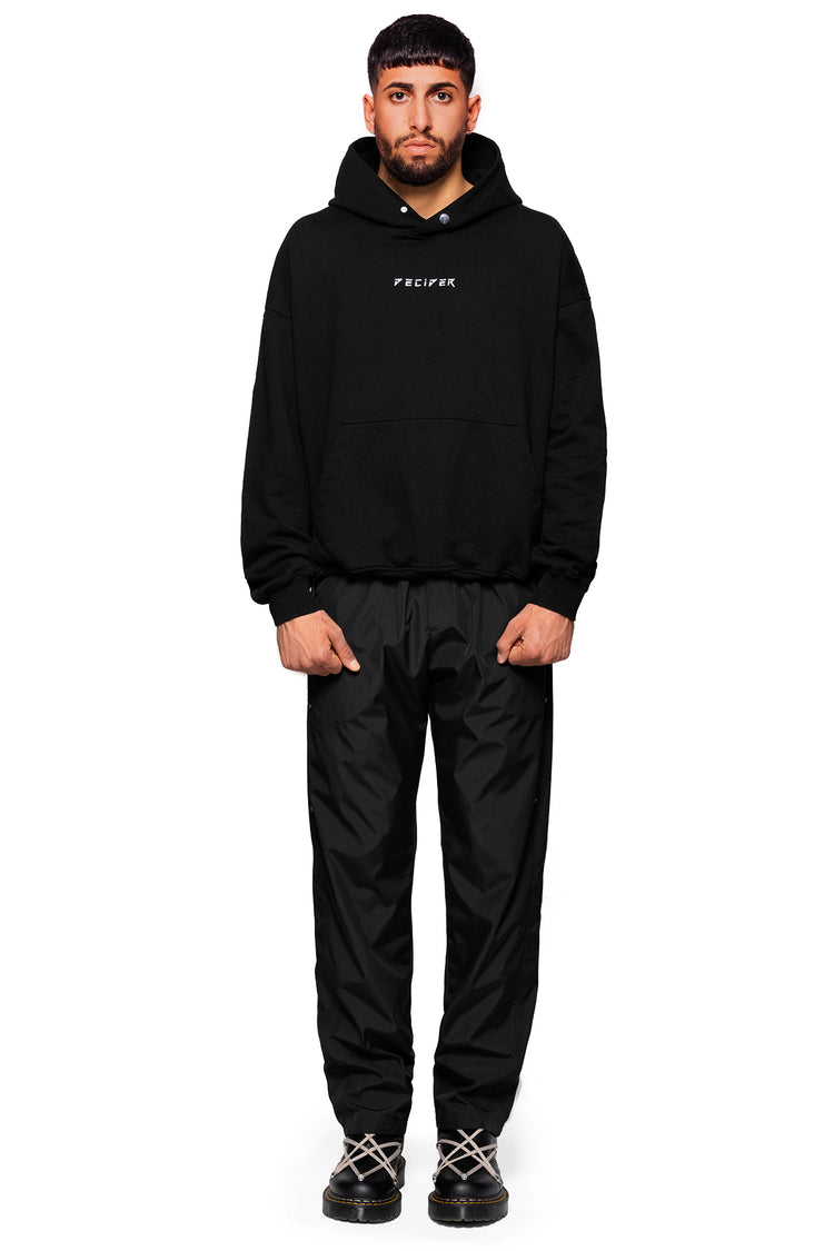 Komfortabler Oversize Hoodie in tiefem Schwarz, ausgestattet mit einer geräumigen Kapuze und Logo-Detail. Getragen mit einer Trackpants und Stiefeln.